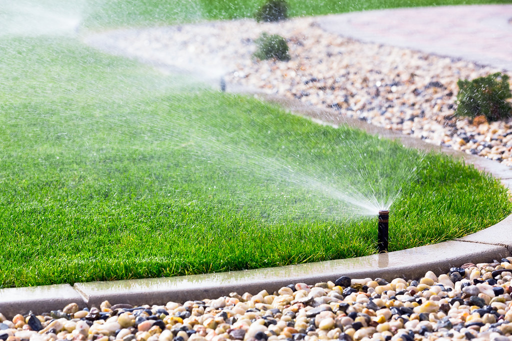 A lawn sprinkler watering grass beside a gravel garden bed and paved edge